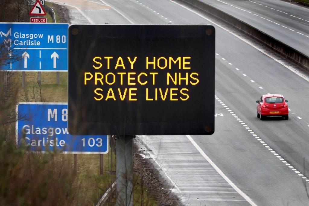 A digital roadside sign above a motorway reading "Stay home, protect NHS, save lives".