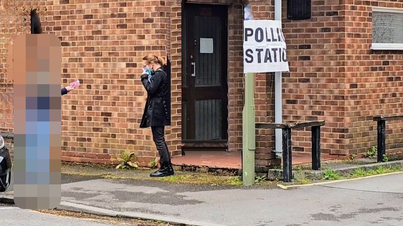Andrea Barrett, wearing a mask, stands outside a polling station while a polling station worker, whose image is blurred, holds out a document.