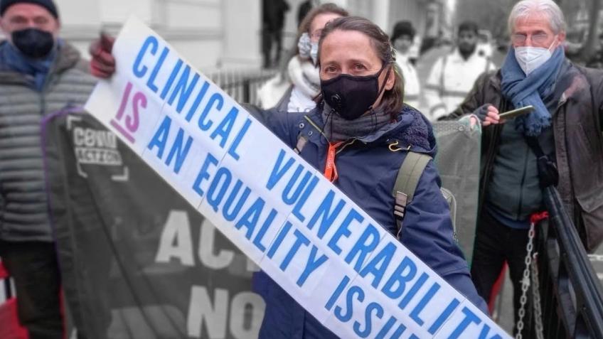 A masked campaigner at a march, beside a banner reading "Clinical vulnerability is an equality issue".