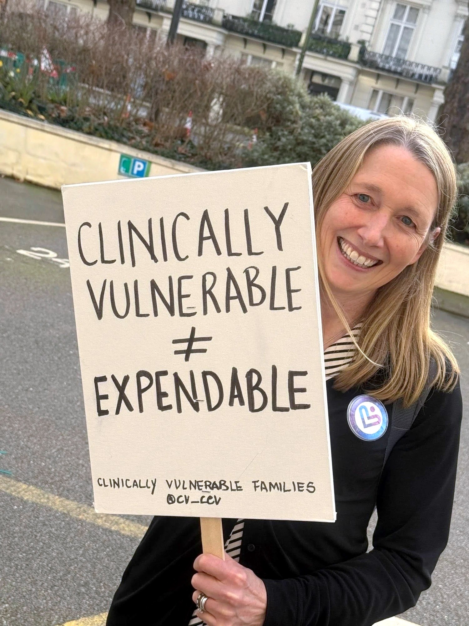 A smiling campaigner wearing a CVF badge holds a placard reading "Clinically vulnerable ≠ expendable".