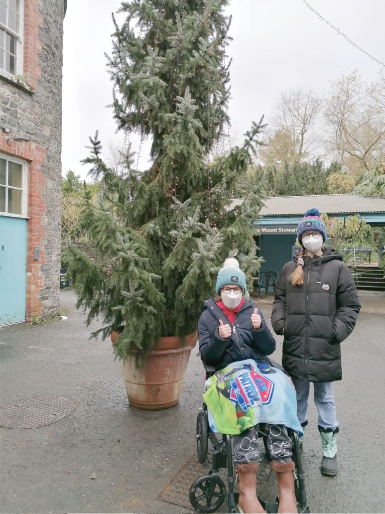 Two people in masks and winter hats beside a large potted Christmas tree outdoors; one is in a wheelchair.