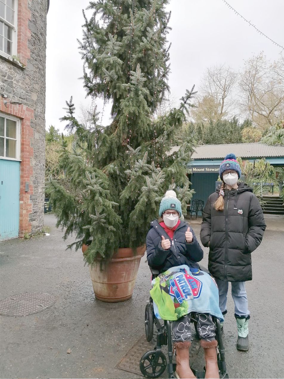 Two people in masks and winter hats beside a large potted Christmas tree outdoors; one is in a wheelchair.