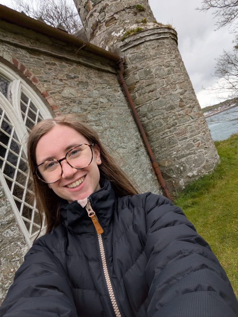 Smiling young woman in glasses and a dark puffer jacket takes a selfie outside a stone building