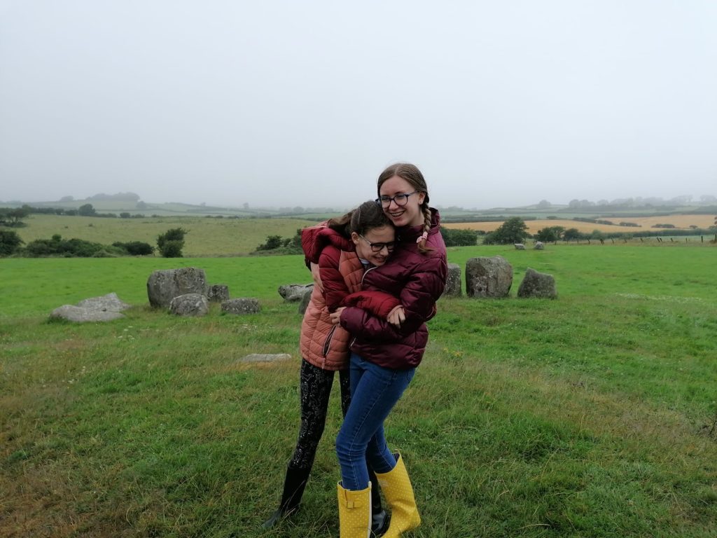 Two young women hug in a green field beside a stone circle on a grey day