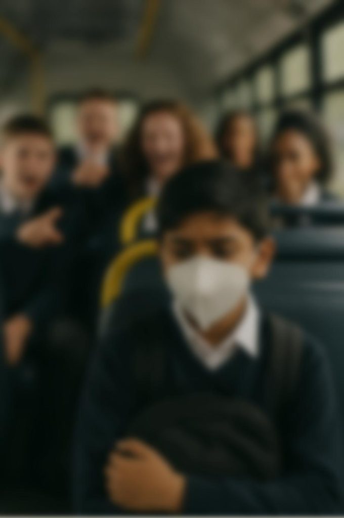 A pupil in school uniform wearing a white mask sits on a school bus while other pupils behind him laugh and point.