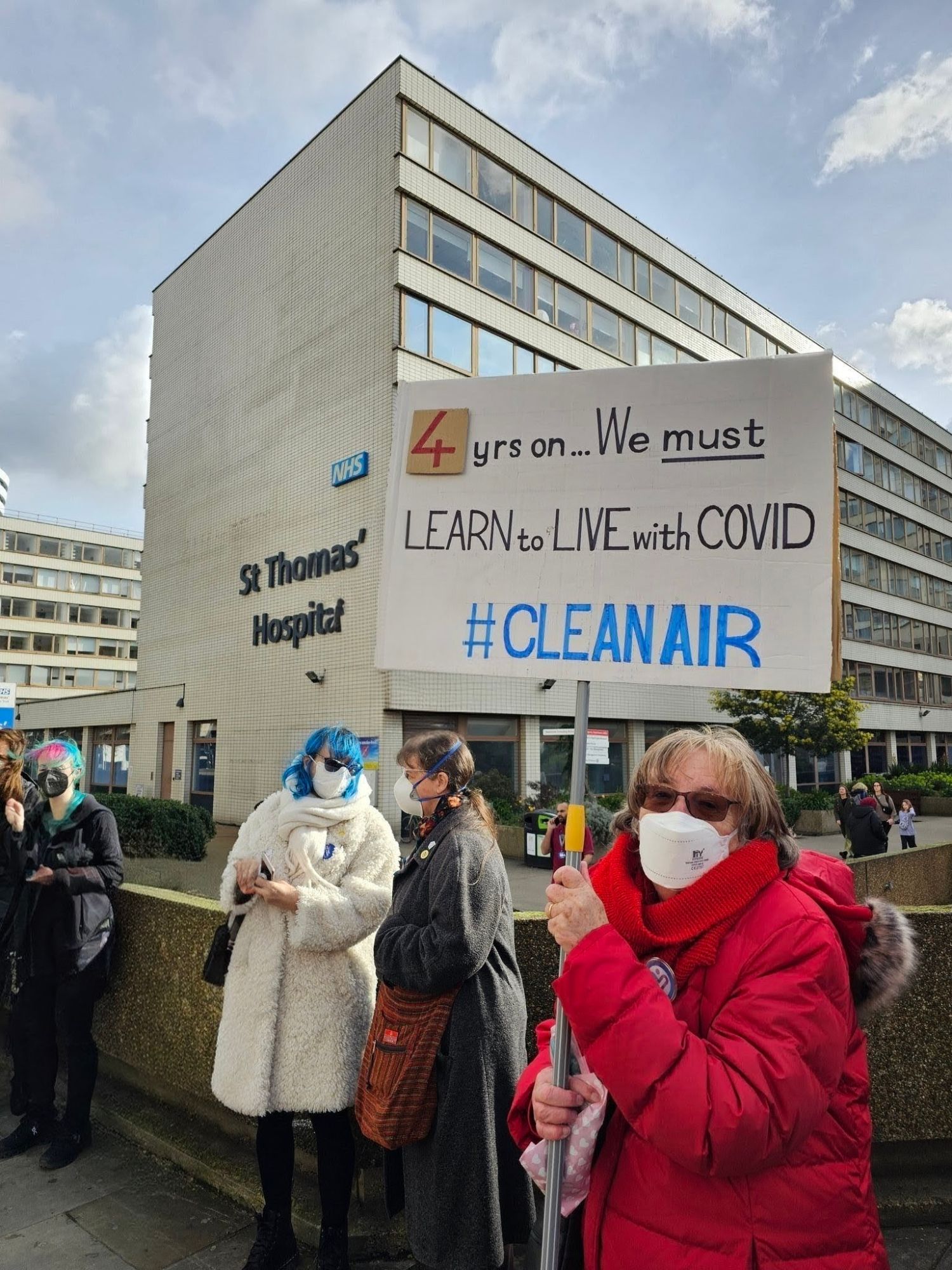 Masked campaigners outside St Thomas’ Hospital in London; one holds a placard reading “4 yrs on… We must learn to live with Covid #CleanAir”.